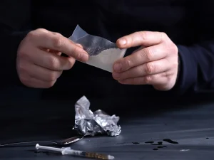 Hands holding a small plastic bag of white powder with drug paraphernalia on a table