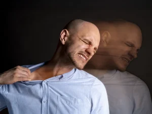 A man grimaces while holding his shoulder, with a blurred double-exposure effect suggesting mental distress