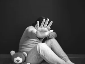 Black and white photo of a child sitting on the floor with hand raised toward the camera, symbolizing a plea to stop abuse and protect victims