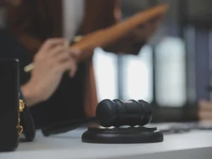 Wooden judge’s gavel resting on a bench in a courtroom with a female judge blurred in the background