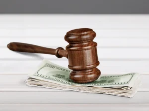 Wooden judge’s gavel resting on a stack of hundred-dollar bills on a white table, symbolizing financial restitution in a criminal case.
