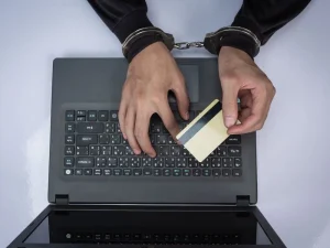 Handcuffed person typing on laptop with credit card, representing cyber fraud charges and imprisonment risk